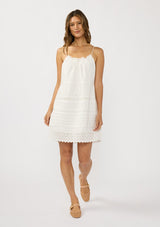 [Color: White] A front facing image of a brunette model wearing a white bohemian sleeveless mini dress with eyelet trims, pintucks, and a ruched neckline with ruffle trims.