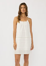 [Color: White] A front facing image of a brunette model wearing a white bohemian sleeveless mini dress with eyelet trims, pintucks, and a ruched neckline with ruffle trims.