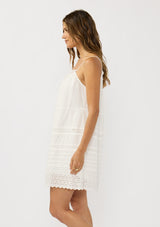 [Color: White] A side facing image of a brunette model wearing a white bohemian sleeveless mini dress with eyelet trims, pintucks, and a ruched neckline with ruffle trims.