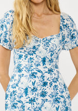 [Color: Ivory/Navy] A detail image of a blonde model wearing a blue and white floral mini dress with short puff sleeves, a sweetheart neckline, side pockets, and a smocked back.