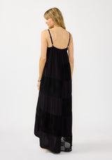 [Color: Black] A back facing image of a blonde model wearing a black bohemian maxi dress with a patchwork lace, sweetheart neckline, tie front detail, and a flowy tiered silhouette. 