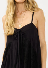 [Color: Black] A detail image of a blonde model wearing a black bohemian maxi dress with a patchwork lace, sweetheart neckline, tie front detail, and a flowy tiered silhouette. 