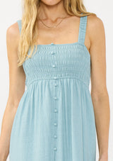 [Color: Aqua Blue] A detail image of a blonde model wearing a light blue midi dress with a smocked bust, button front, and ruffle tiered  hemline. 