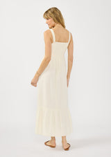 [Color: Tea] A back facing image of a blonde model wearing a light beige sleeveless maxi dress with lace trims, a square neckline, and a smocked back. 