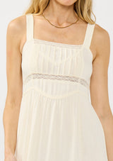 [Color: Tea] A detail image of a blonde model wearing a light beige sleeveless maxi dress with lace trims, a square neckline, and a smocked back. 