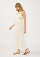 [Color: Tea] A side facing image of a blonde model wearing a light beige sleeveless maxi dress with lace trims, a square neckline, and a smocked back. 