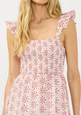 [Color: Paprika/White] A detail image of a blonde model wearing a red floral midi dress with eyelets. Featuring a smocked bust, elastic straps with ruffle detail, and a tie back detail.