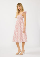 [Color: Paprika/White] A side facing image of a blonde model wearing a red floral midi dress with eyelets. Featuring a smocked bust, elastic straps with ruffle detail, and a tie back detail.