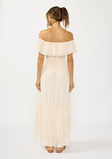 [Color: Tea Dye] A back facing image of a woman in a light beige maxi dress with an off shoulder design, lace trims, a smocked waist, and side pockets.