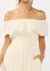 [Color: Tea Dye] A detail image of a woman in a light beige maxi dress with an off shoulder design, lace trims, a smocked waist, and side pockets.