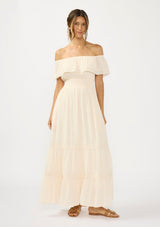 [Color: Tea Dye] A front facing image of a woman in a light beige maxi dress with an off shoulder design, lace trims, a smocked waist, and side pockets.