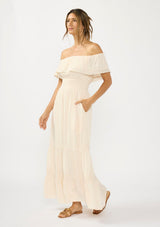 [Color: Tea Dye] A side facing image of a woman in a light beige maxi dress with an off shoulder design, lace trims, a smocked waist, and side pockets.