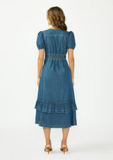 [Color: Denim] A back facing image of a brunette model wearing a denim blue midi dress with a v neckline, short puff sleeves, a button front, and a tiered hem with raw edges. Paired with closed toe suede heels. 