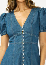 [Color: Denim] A detail image of a brunette model wearing a denim blue midi dress with a v neckline, short puff sleeves, a button front, and a tiered hem with raw edges. Paired with closed toe suede heels. 