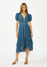 [Color: Denim] A front facing image of a brunette model wearing a denim blue midi dress with a v neckline, short puff sleeves, a button front, and a tiered hem with raw edges. Paired with closed toe suede heels. 