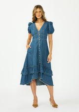 [Color: Denim] A front facing image of a brunette model wearing a denim blue midi dress with a v neckline, short puff sleeves, a button front, and a tiered hem with raw edges. Paired with closed toe suede heels. 
