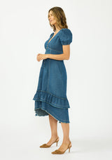[Color: Denim] A side facing image of a brunette model wearing a denim blue midi dress with a v neckline, short puff sleeves, a button front, and a tiered hem with raw edges. Paired with closed toe suede heels. 