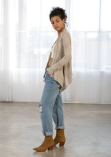 [Color: Warm Grey] A side facing image of a brunette model wearing a pointelle knit cardigan with metallic thread details, a waterfall front, and long cozy sleeves. 