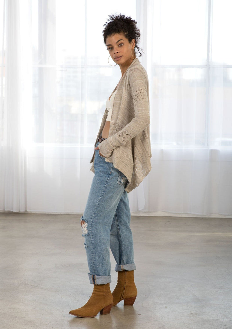 [Color: Warm Grey] A side facing image of a brunette model wearing a pointelle knit cardigan with metallic thread details, a waterfall front, and long cozy sleeves. 