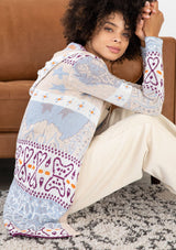 [Color: Sky/Plum] A sitting image of a brunette model wearing a vibrant boho patterned knit cardigan with long cozy sleeves, open front, and hoodie. An effortless statement sweater for easy layering.  