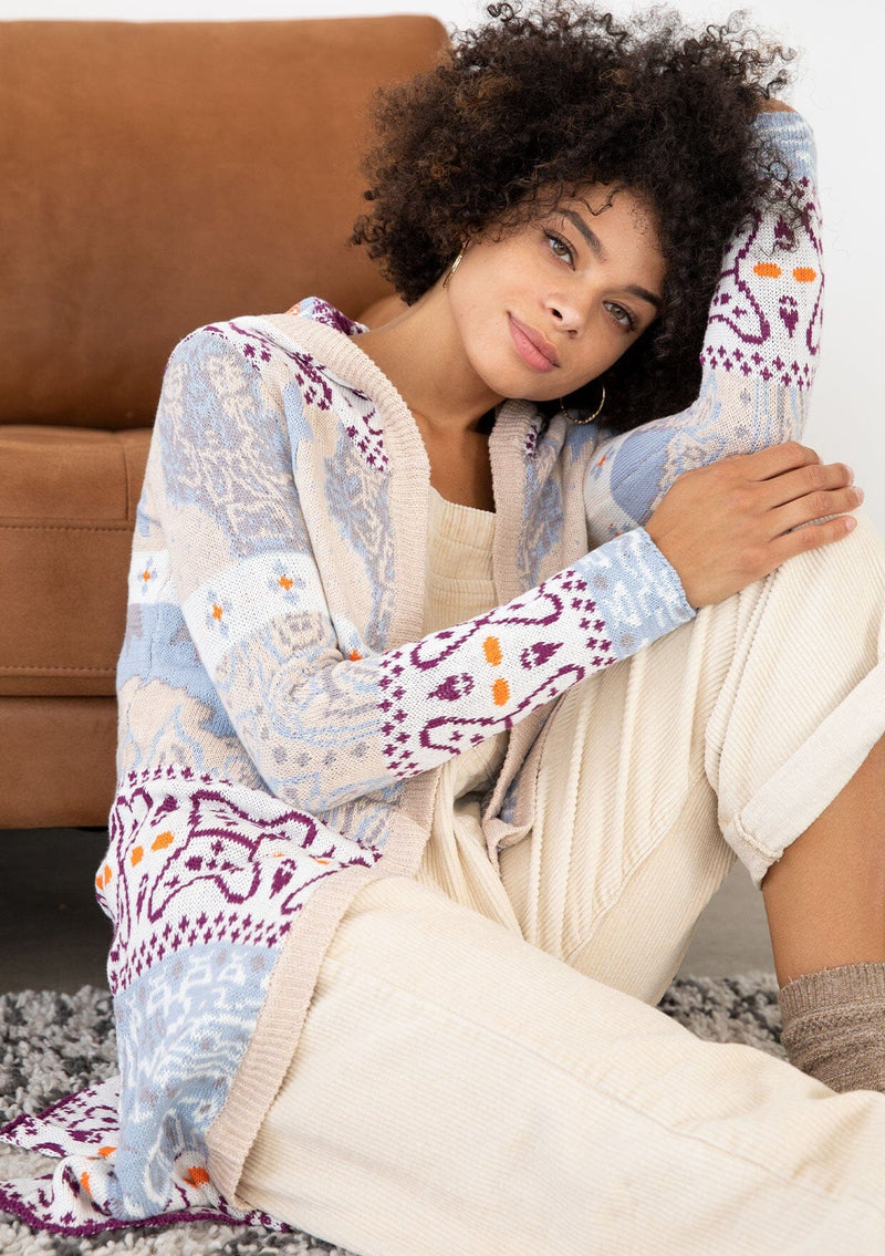 [Color: Sky/Plum] A sitting image of a brunette model wearing a vibrant boho patterned knit cardigan with long cozy sleeves, open front, and hoodie. An effortless statement sweater for easy layering.  