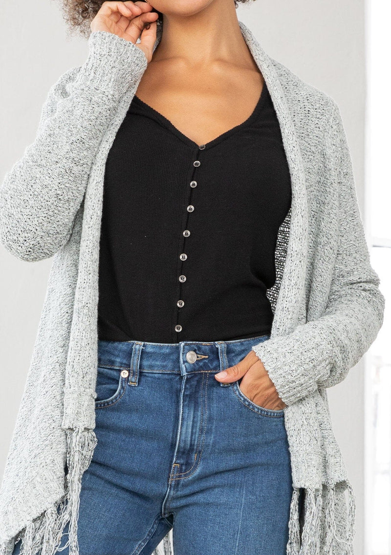 [Color: Grey/Ivory] A detail image of a marled knit cardigan with a shawl waterfall collar with a fringe hem. A boho chic cardigan for the fall season. 