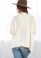 [Color: Cream] A back facing image of a brunette model wearing a cozy ivory open knit cardigan with a shawl collar, cable knit details, and ribbed trims. A casual fall cardigan for any occasion. 