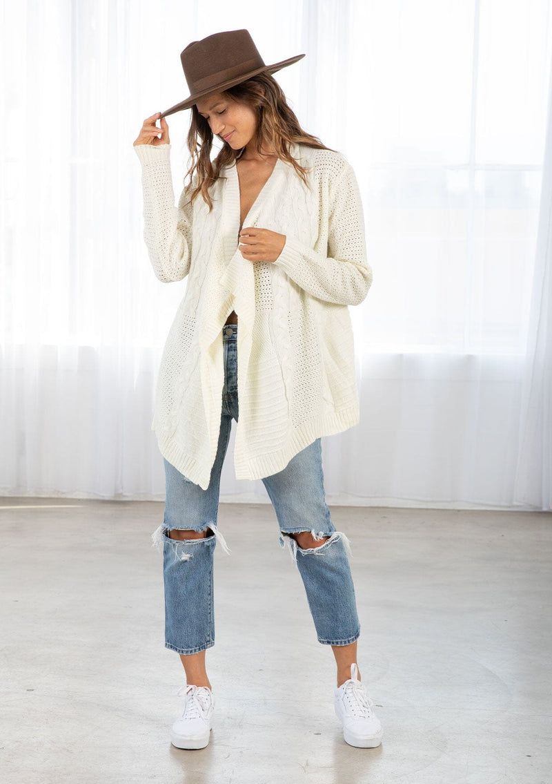 [Color: Cream] A front facing image of a brunette model wearing a cozy ivory open knit cardigan with a shawl collar, cable knit details, and ribbed trims. A casual fall cardigan for any occasion. 