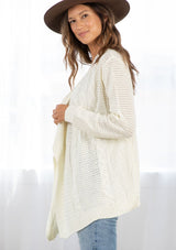 [Color: Cream] A side facing image of a brunette model wearing a cozy ivory open knit cardigan with a shawl collar, cable knit details, and ribbed trims. A casual fall cardigan for any occasion. 