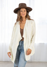 [Color: Cream] A front facing image of a brunette model wearing a cozy ivory open knit cardigan with a shawl collar, cable knit details, and ribbed trims. A casual fall cardigan for any occasion. 