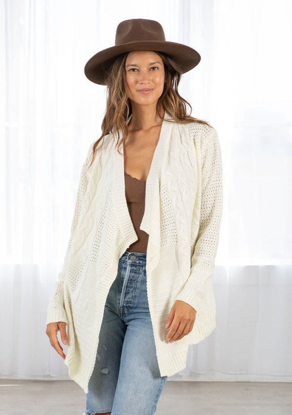 [Color: Cream] A front facing image of a brunette model wearing a cozy ivory open knit cardigan with a shawl collar, cable knit details, and ribbed trims. A casual fall cardigan for any occasion. 