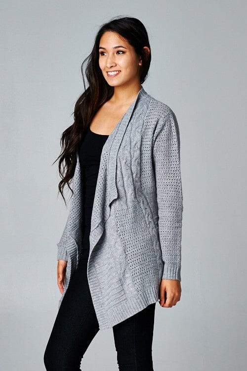 [Color: Grey] A side facing image of a brunette model wearing a cozy grey open knit cardigan with a shawl collar, cable knit details, and ribbed trims. A casual fall cardigan for any occasion. 