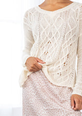 [Color: Vanilla] A detail image of a brunette model wearing a lightweight cable knit pullover with open knit details. Featuring a classic round neckline, dropped shoulders, long sleeves, and a shirttail hem. Laid-back boho style for casual outings. 