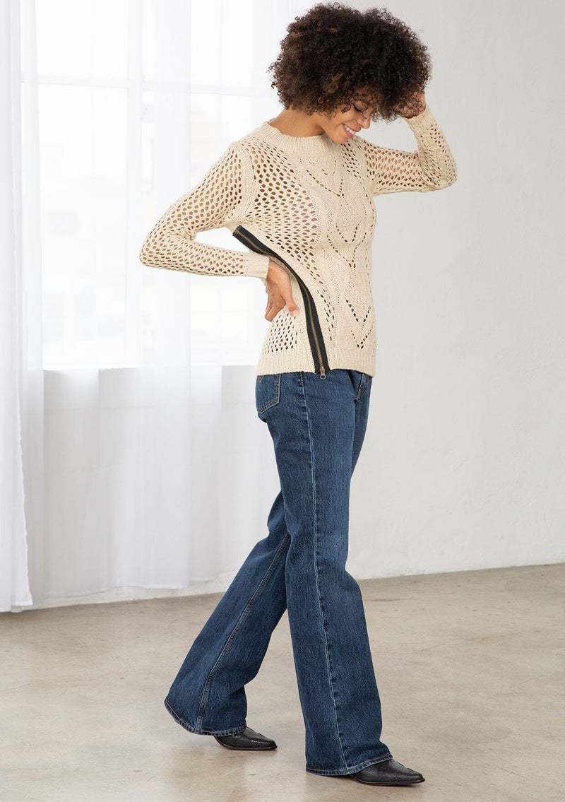 [Color: Natural] An image of a brunette model wearing a beige crochet pullover sweater with a side zipper accent. Featuring a crew neckline, long sleeves, and rib trim. 
