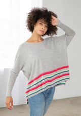 [Color: Silver/Multi] A side facing image of a brunette model wearing a grey pullover with a vibrant stripe accent. Featuring a relaxed fit, long dolman sleeves with tapered  finish, and a flattering round neckline. 