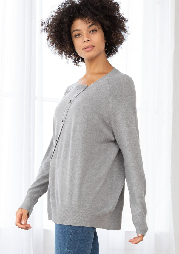 [Color: Heather Grey] A side facing image of a brunette model wearing a grey pullover button front cardigan with an asymmetrical design and long relaxed cozy sleeves. 