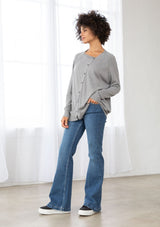 [Color: Heather Grey] A side facing image of a brunette model wearing a grey pullover button front cardigan with an asymmetrical design and long relaxed cozy sleeves. 