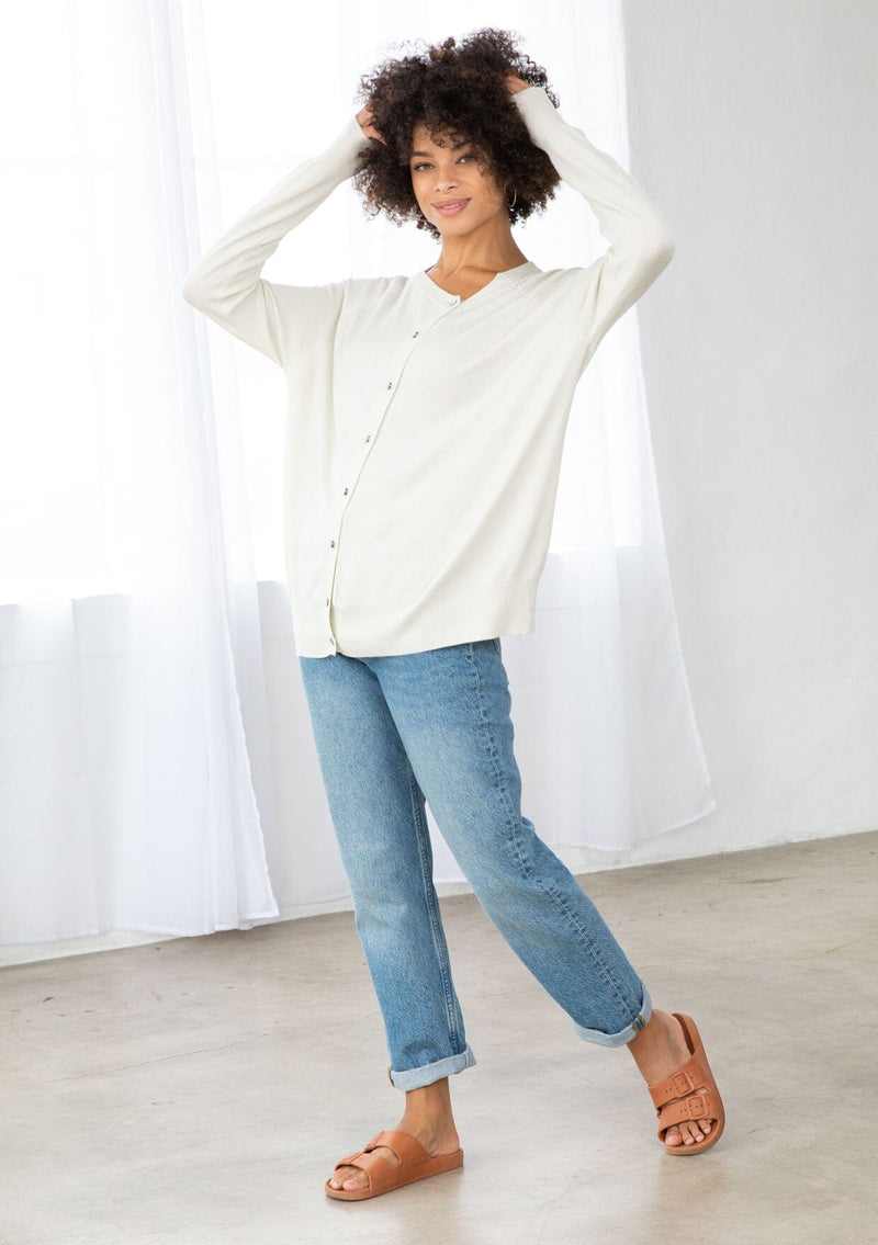 [Color: Bone] A front facing image of a brunette model wearing a white pullover button front cardigan with an asymmetrical design and long relaxed cozy sleeves. 