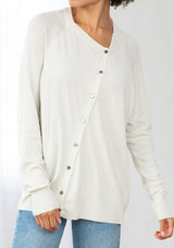 [Color: Bone] A front facing image of a brunette model wearing a white pullover button front cardigan with an asymmetrical design and long relaxed cozy sleeves. 