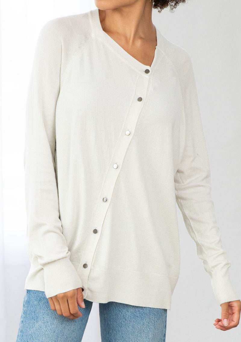 [Color: Bone] A front facing image of a brunette model wearing a white pullover button front cardigan with an asymmetrical design and long relaxed cozy sleeves. 