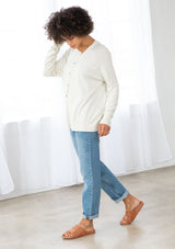 [Color: Bone] A side facing image of a brunette model wearing a white pullover button front cardigan with an asymmetrical design and long relaxed cozy sleeves. 