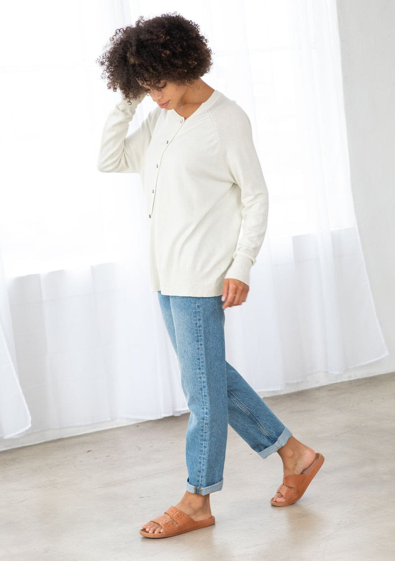 [Color: Bone] A side facing image of a brunette model wearing a white pullover button front cardigan with an asymmetrical design and long relaxed cozy sleeves. 
