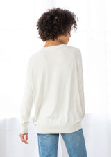 [Color: Bone] A back facing image of a brunette model wearing a white pullover button front cardigan with an asymmetrical design and long relaxed cozy sleeves. 