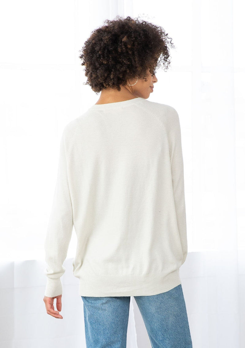 [Color: Bone] A back facing image of a brunette model wearing a white pullover button front cardigan with an asymmetrical design and long relaxed cozy sleeves. 