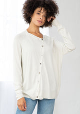 [Color: Bone] A front facing image of a brunette model wearing a white pullover button front cardigan with an asymmetrical design and long relaxed cozy sleeves. 