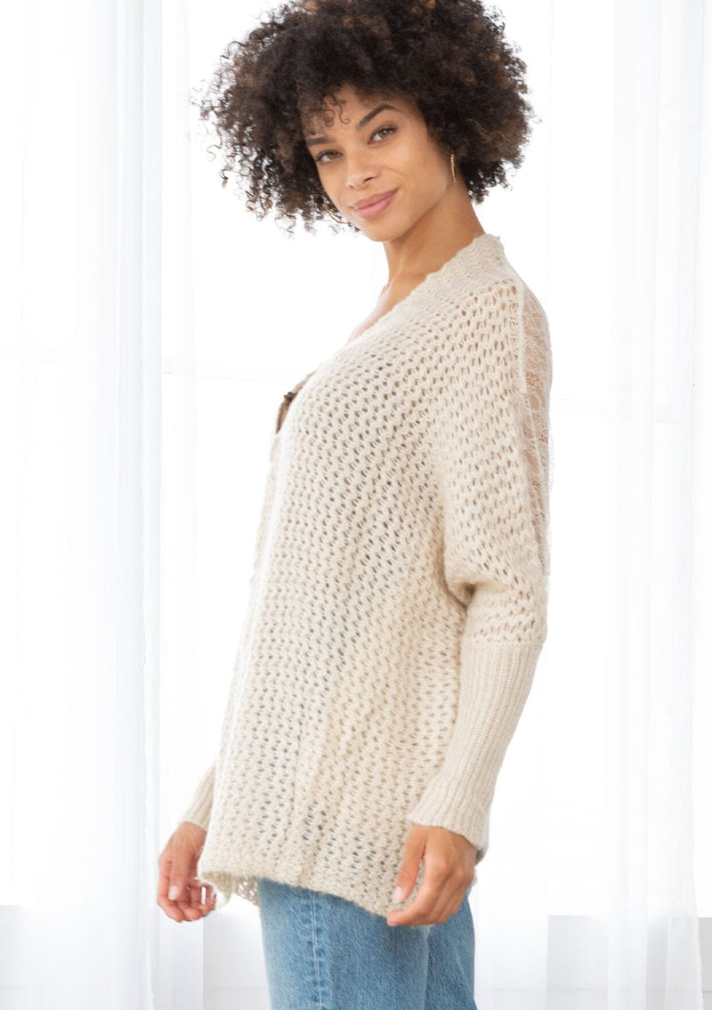 [Color: Oatmeal] A side facing image of a brunette model wearing a knit and lace beige cardigan with a hook and eye closure, batwing sleeves, and slim sleeve finish. A cozy and effortless cardigan for the fall season. 