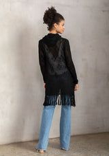 [Color: Black] A back facing image of a brunette model wearing a crochet diamond pattern cardigan with a drawstring hoodie, fringe hemline, and an open front. A classic boho layer for any season. 