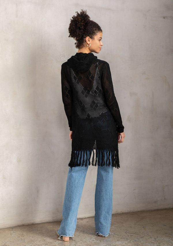 [Color: Black] A back facing image of a brunette model wearing a crochet diamond pattern cardigan with a drawstring hoodie, fringe hemline, and an open front. A classic boho layer for any season. 