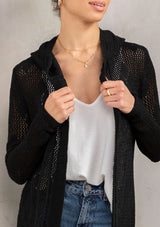 [Color: Black] A detail image of a brunette model wearing a crochet diamond pattern cardigan with a drawstring hoodie, fringe hemline, and an open front. A classic boho layer for any season. 
