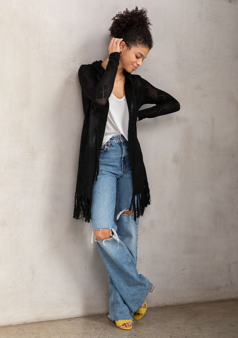 [Color: Black] A front facing image of a brunette model wearing a crochet diamond pattern cardigan with a drawstring hoodie, fringe hemline, and an open front. A classic boho layer for any season. 